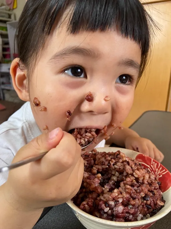 茶碗に山盛りの喝米を、スプーンで口いっぱいに頬張る子ども。顔にお米の粒がついている